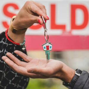 Close-up of hands exchanging house key with colorful SOLD sign in the background.