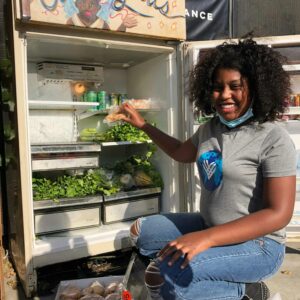 Smiling woman stocks a community fridge with fresh greens and groceries outdoors.